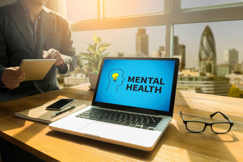 A laptop on a desk displays the words MENTAL HEALTH with a head and brain graphic. Nearby are glasses, a notebook, a pen, a phone, and a person using a tablet, with a city skyline visible through the window.