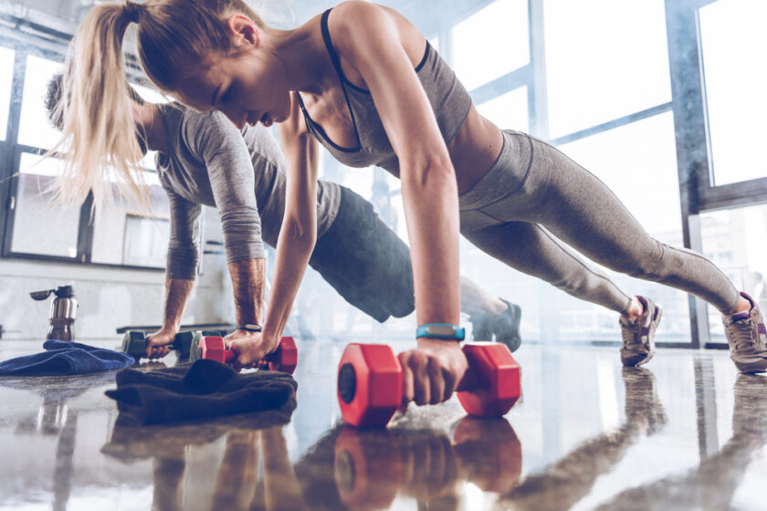 Two people in athletic wear perform push-ups using red dumbbells in a bright gym with large windows. They appear focused, exercising side by side on a shiny floor with workout towels nearby.