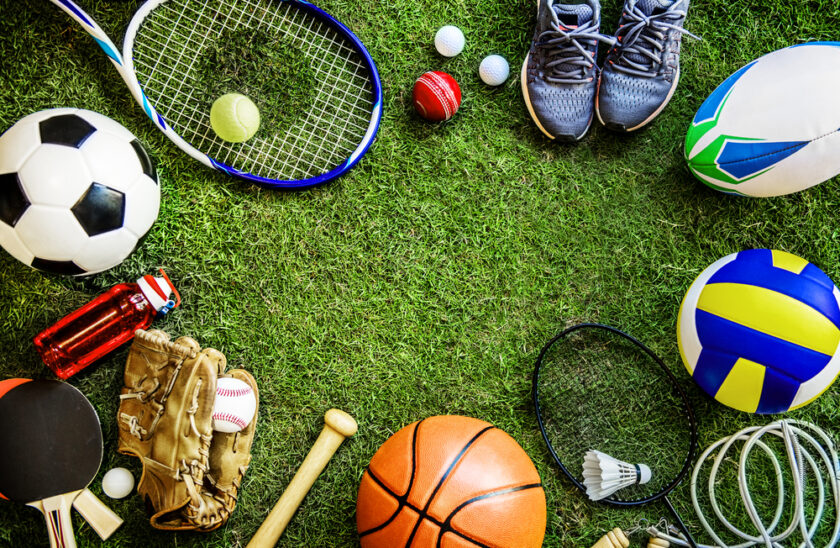 Various sports equipment, including a soccer ball, tennis racket, baseball glove, basketball, volleyball, table tennis paddle, running shoes, water bottles, and more, arranged in a circle on green grass.