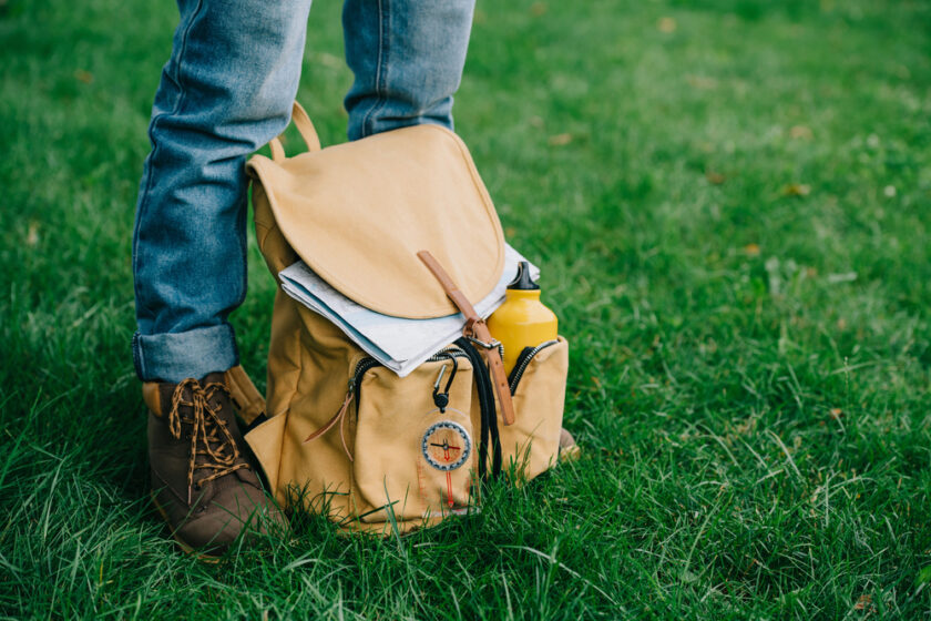A person wearing brown boots and blue jeans stands on green grass next to a yellow backpack holding a notebook and a yellow water bottle.