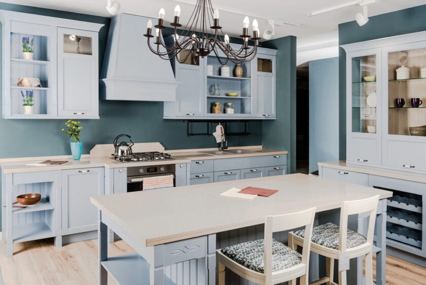 A spacious kitchen with light blue cabinets, a central island with three chairs, open shelving, a gas stove, and a black chandelier. Light wood flooring and teal walls complement the airy, modern design.