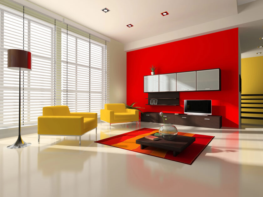 A modern living room with a bold red accent wall, two yellow armchairs, a low coffee table with a glass bowl, a red and orange rug, floor lamp, large windows with blinds, and floating shelves with a TV.