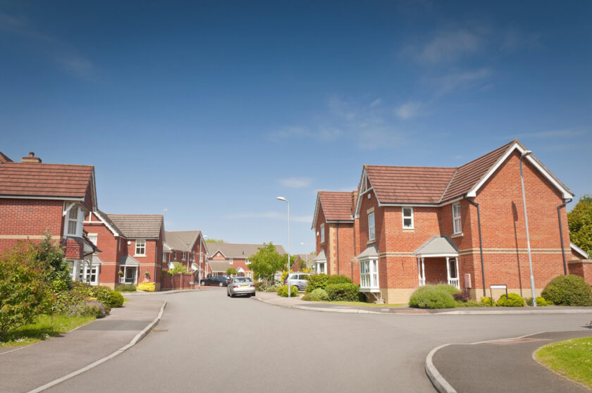 A quiet suburban street with modern red-brick houses, neat gardens, and parked cars under a clear blue sky on a sunny day.