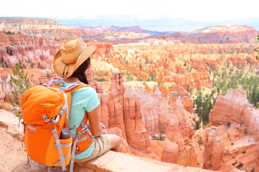 A woman wearing a hat and an orange backpack sits on a stone ledge, overlooking the colorful rock formations and canyons of Bryce Canyon National Park under a bright sky.