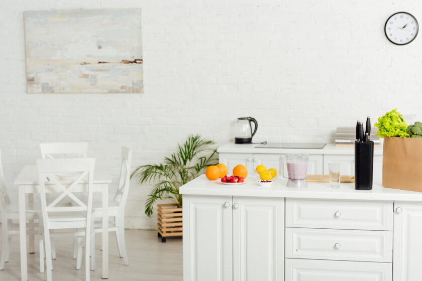 Bright kitchen with white cabinets and a dining table. On the counter are oranges, strawberries, a smoothie, a glass, a black knife block, and a paper bag with leafy greens. A clock and artwork hang on the white wall.
