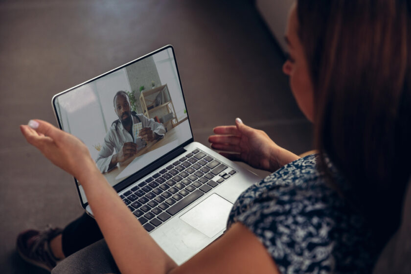 A woman sits and gestures while having a video call on her laptop with a man holding playing cards. The mans background shows shelves and a plant, indicating they are in different locations.
