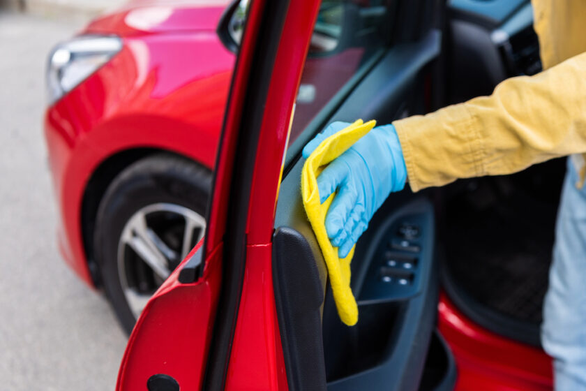 A person wearing blue gloves uses a yellow cloth to clean the interior door panel of a red car, with the door open and the exterior of the car visible in the background.