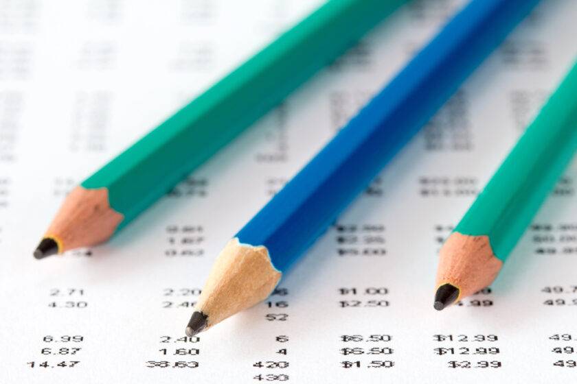 Three colored pencils (green, blue, and teal) lie diagonally on a sheet of paper covered with rows of printed numbers, suggesting financial data or calculations.
