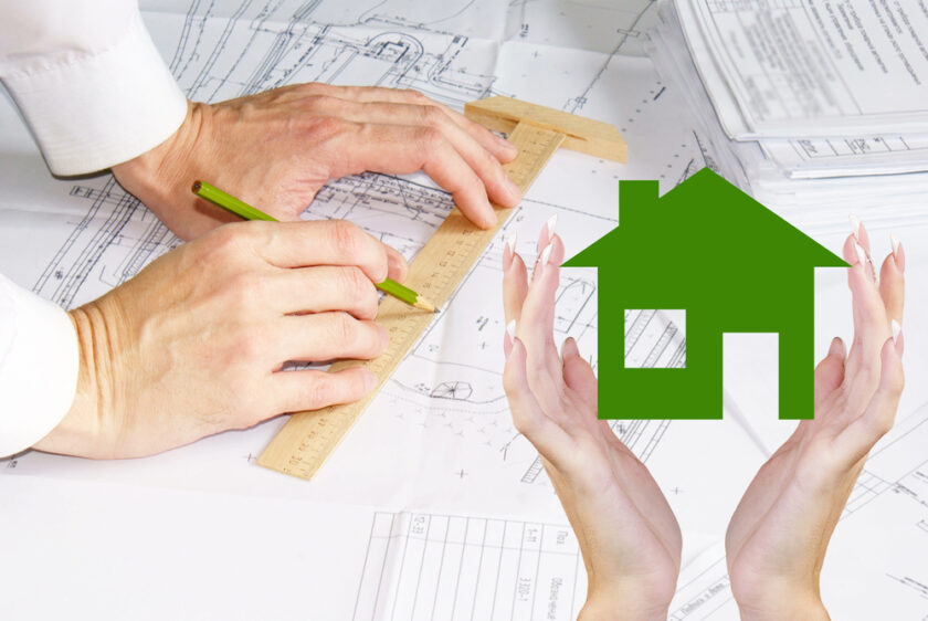 A person draws on architectural blueprints with a ruler and pencil. In the foreground, two hands frame a green house icon, symbolizing home building, planning, or real estate concepts.