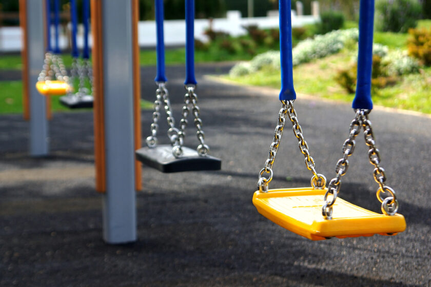 Close-up of empty swings on a playground, with yellow and black seats hanging from blue chains over a rubberized ground surface. Bushes and greenery are visible in the blurred background.