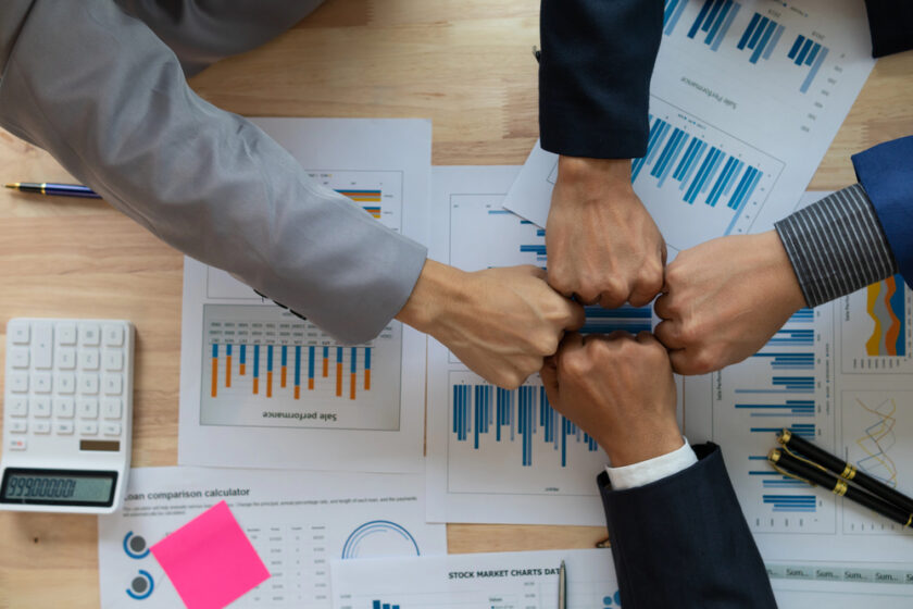 Four people in business attire perform a group fist bump over a table covered with financial charts, graphs, a calculator, pen, and documents, symbolizing teamwork and collaboration in a professional setting.