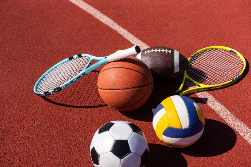Five sports items—two tennis rackets, a basketball, an American football, a volleyball, and a soccer ball—are placed together on a red running track near a white line.