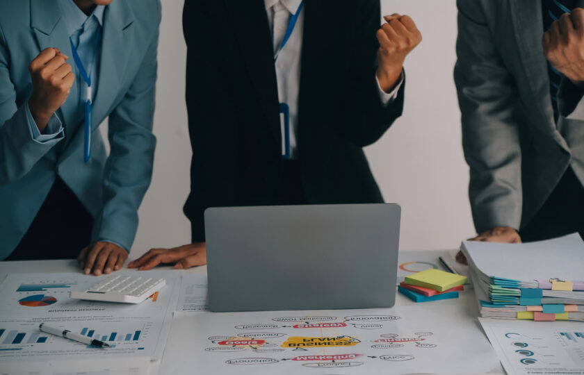 Three businesspeople stand around a desk with charts, documents, and a laptop, raising their fists in a gesture of success or motivation, suggesting teamwork and achievement in a professional setting.