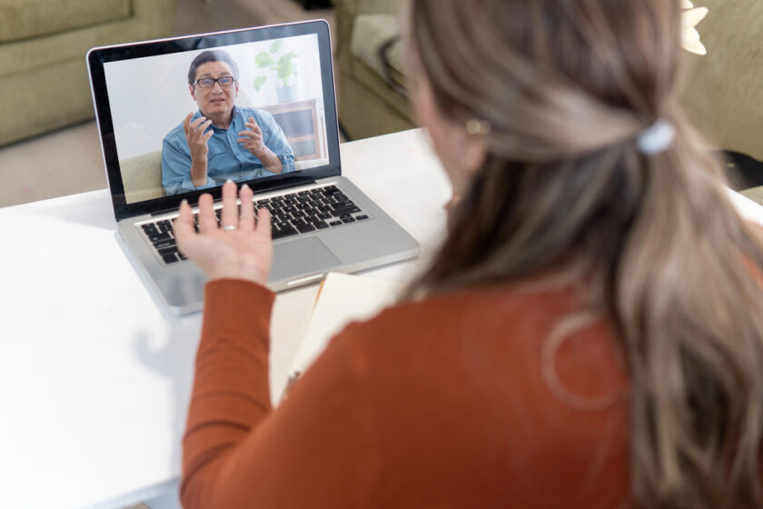 A woman with light brown hair has a video call on her laptop with a man wearing glasses and a blue shirt, who gestures with his hands. She is seated at a white table with a notebook in front of her.