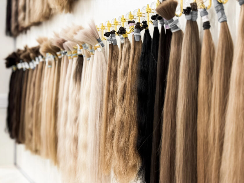 Bundles of hair extensions in various shades, from light blonde to dark brown, are neatly displayed hanging on a wall, organized by color and texture.