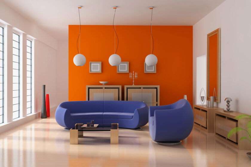Modern living room with a bright orange accent wall, three spherical pendant lights, blue curved sofas, light wood flooring, and large windows letting in natural light. Minimalist decor with geometric accents.