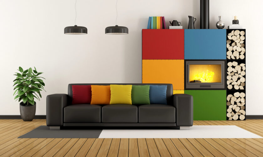 Modern living room with a black sofa featuring colorful cushions, a plant on the left, and a fireplace set in a wall with large red, yellow, blue, and green panels and stacked firewood on the right.