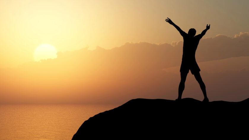 Silhouette of a person standing on a rocky hill with arms raised in triumph, overlooking a calm sea at sunset, with the sun partially hidden by clouds.
