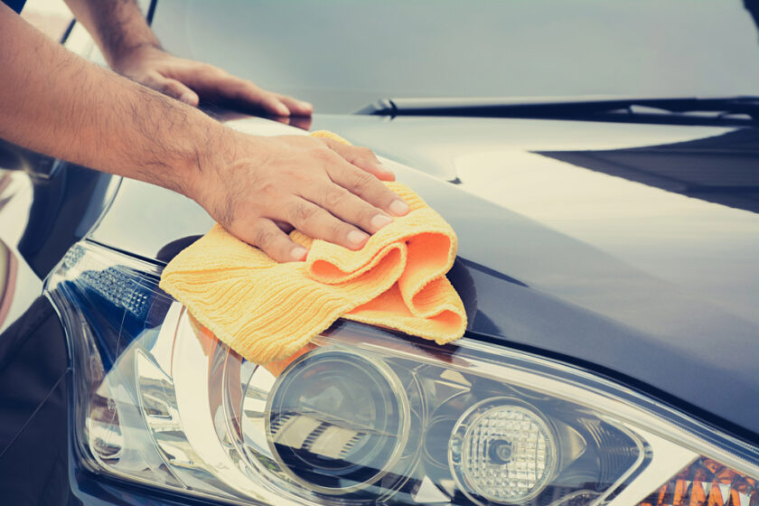 A person’s hands polishing the hood and headlight of a black car with a yellow microfiber cloth.