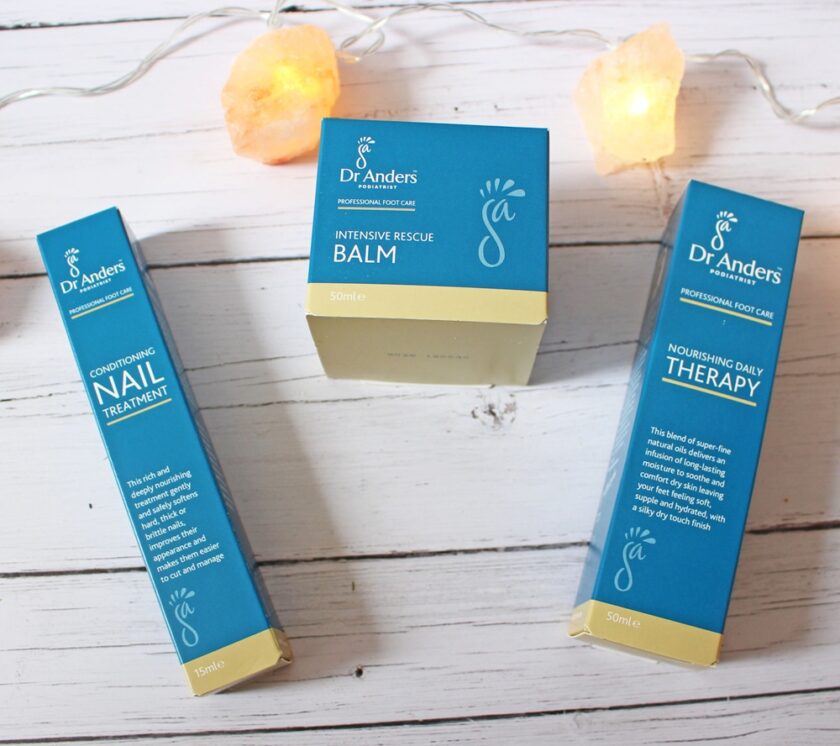 Three Dr. Anders skincare products—Nail Treatment, Intensive Rescue Balm, and Daily Therapy—are arranged on a white wooden surface with string lights and two orange decorative stones in the background.