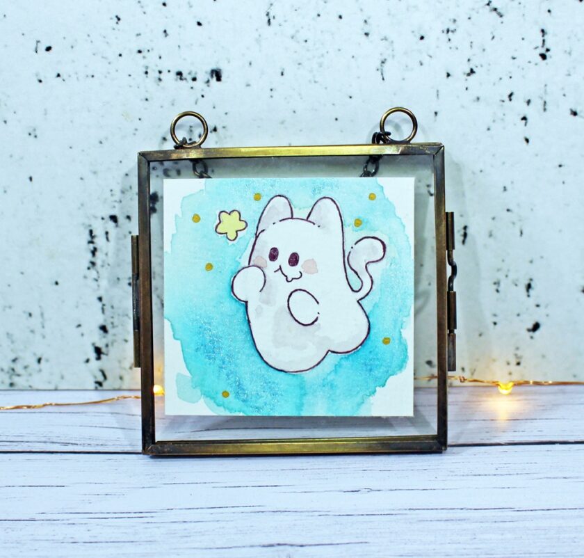 A framed drawing of a cute, chubby cat with a curled tail, surrounded by blue watercolor and yellow dots, hangs in front of a white speckled wall. Tiny fairy lights are visible on the surface below.