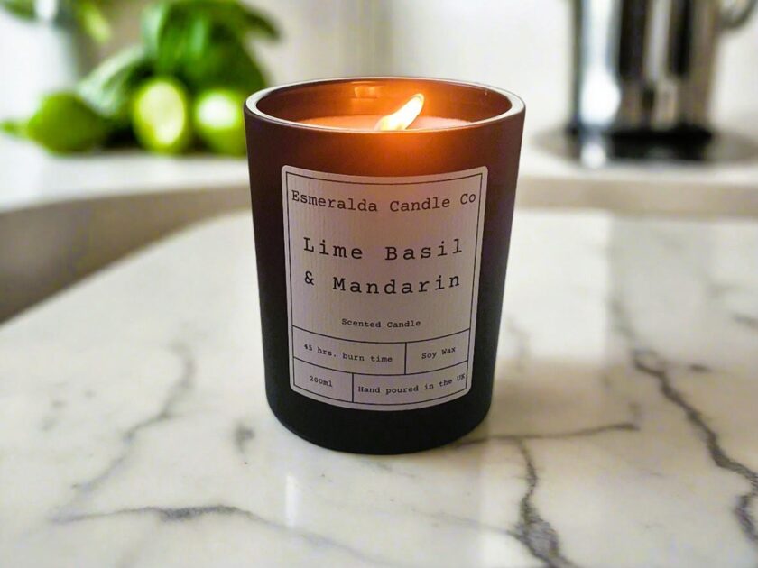 A brown candle labeled Esmerralda Candle Co Lime Basil & Mandarin burns on a white marble surface. The candles label notes a 45 hr burn time, 200ml size, and that it is hand poured in the UK. Out-of-focus limes are in the background.