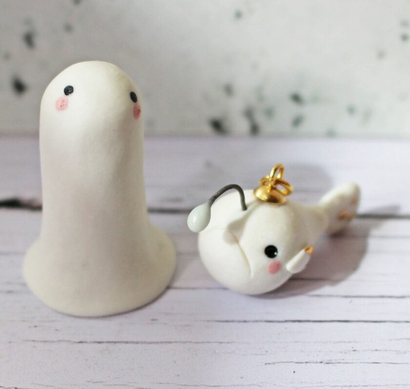 Two small, white, clay figurines with simple black eyes and pink cheeks; one stands upright, while the other lies on its side with a gold loop and wire detail on its head, on a pale wooden surface.