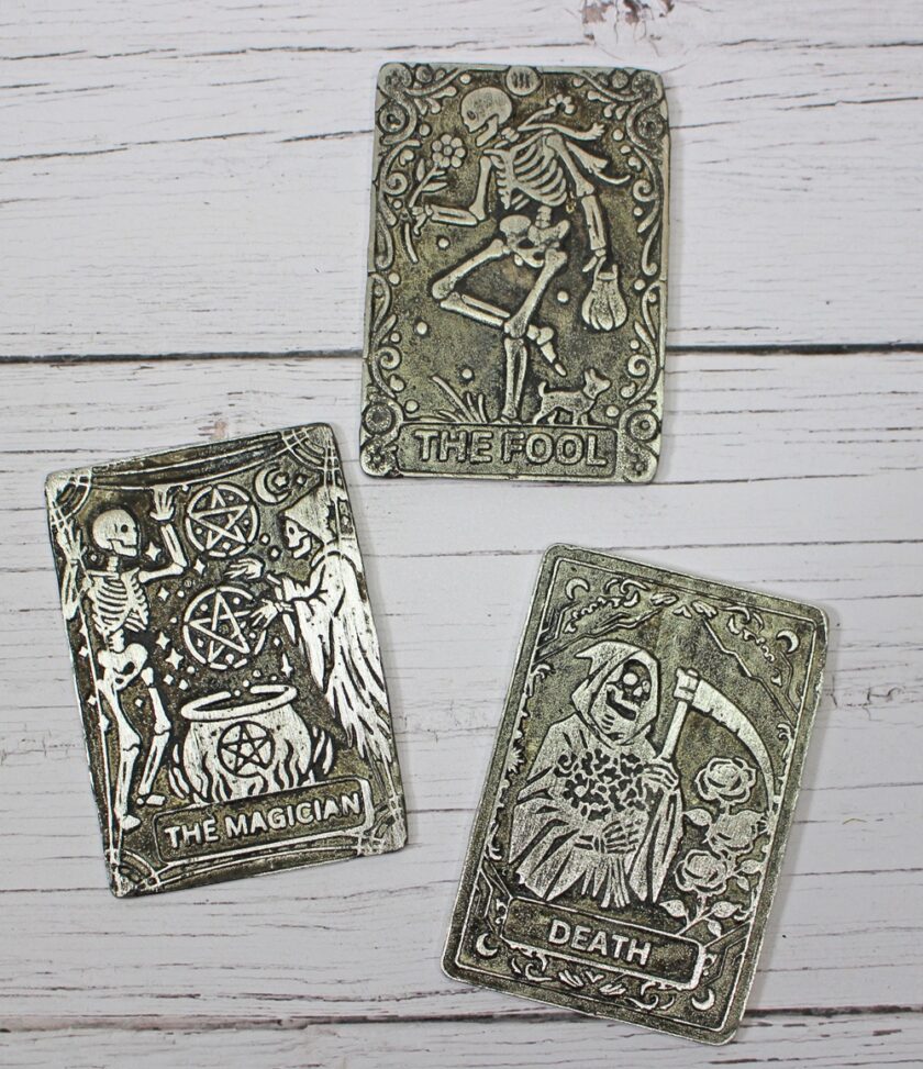 Three metallic tarot cards on a white wooden surface, featuring illustrations for The Fool (skeleton dancing), The Magician (skeleton conjuring symbols), and Death (skeleton with a scythe and rose).