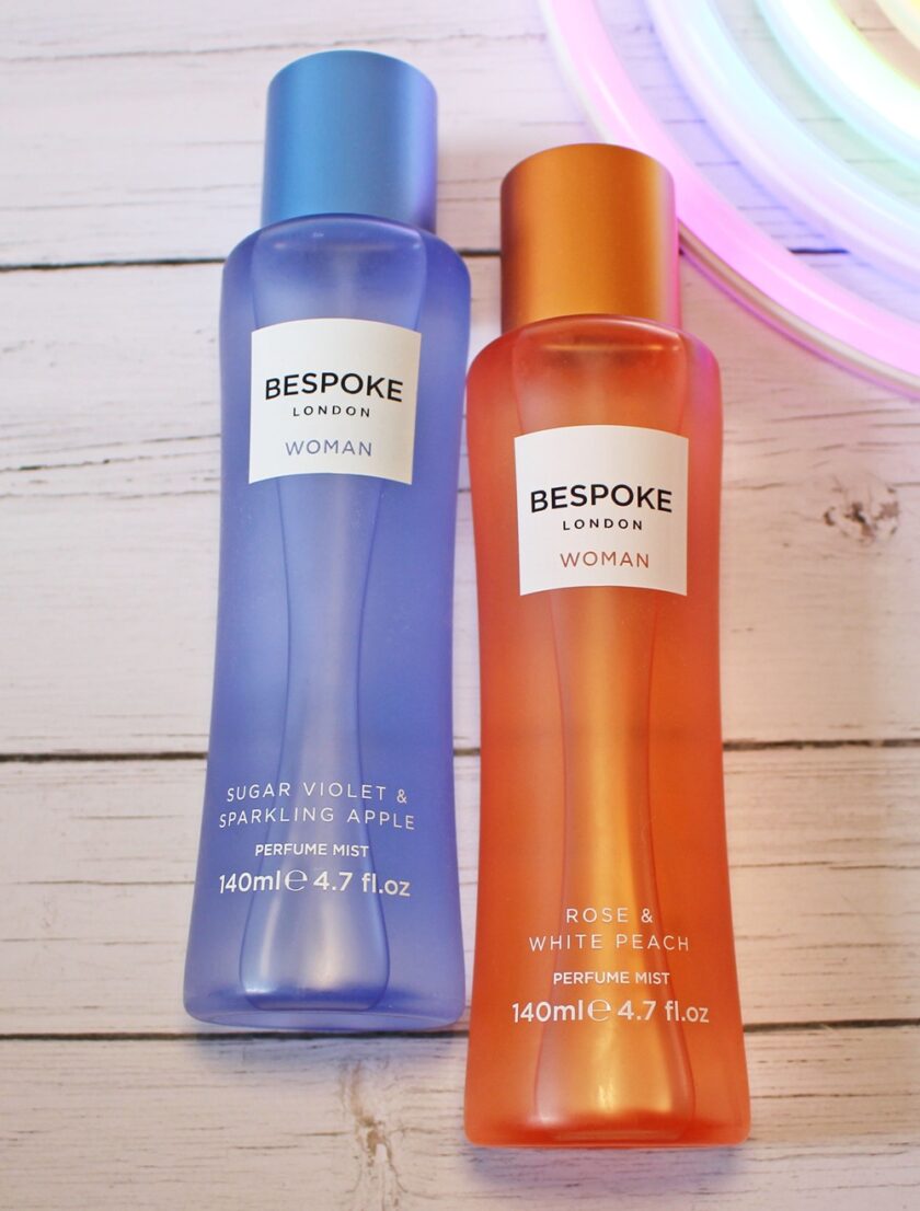 Two Bespoke London perfume mist bottles on a white wooden surface; one is blue labelled Sugar Violet & Sparkling Apple, the other is orange labelled Rose & White Peach. A neon light is visible in the corner.