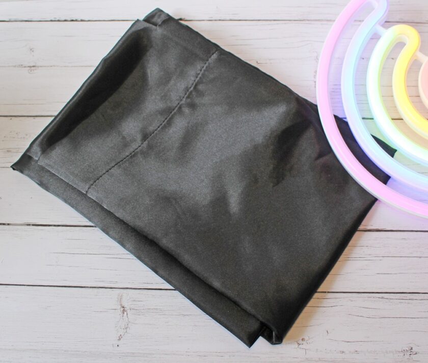 A folded piece of black fabric lies on a light wooden surface next to a partially visible colourful neon rainbow light.