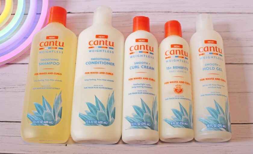 Five Cantu Weightless hair care products for waves and curls, including shampoo, conditioner, curl cream, daily oil, and hold gel, are lined up on a light wooden surface with a colourful object on the side.