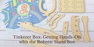 A robotic hand kit with wooden parts and a colorful instruction sheet is laid out on a light wooden surface. Text reads: Tinkerer Box: Getting Hands-On with the Robotic Hand Box.