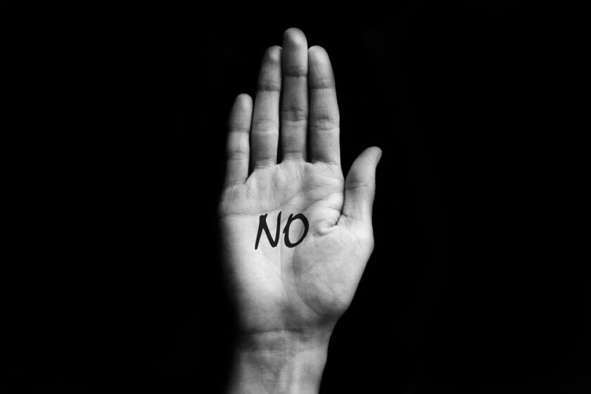 A raised hand with the palm facing forwards against a black background. The word NO is written in bold black letters on the centre of the palm.