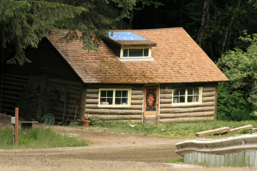 A small log cabin with a shingled roof, an attic window, and a colourful star pattern on the front door sits surrounded by greenery and trees, near a dirt road and stacked logs or fence rails in the foreground.