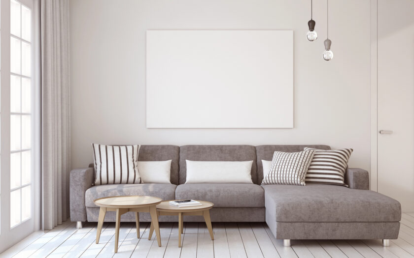 A modern living room with a grey sectional sofa, striped and plain cushions, two round wooden coffee tables, large blank artwork on the wall, hanging light bulbs, and sunlight coming through tall windows with sheer curtains.