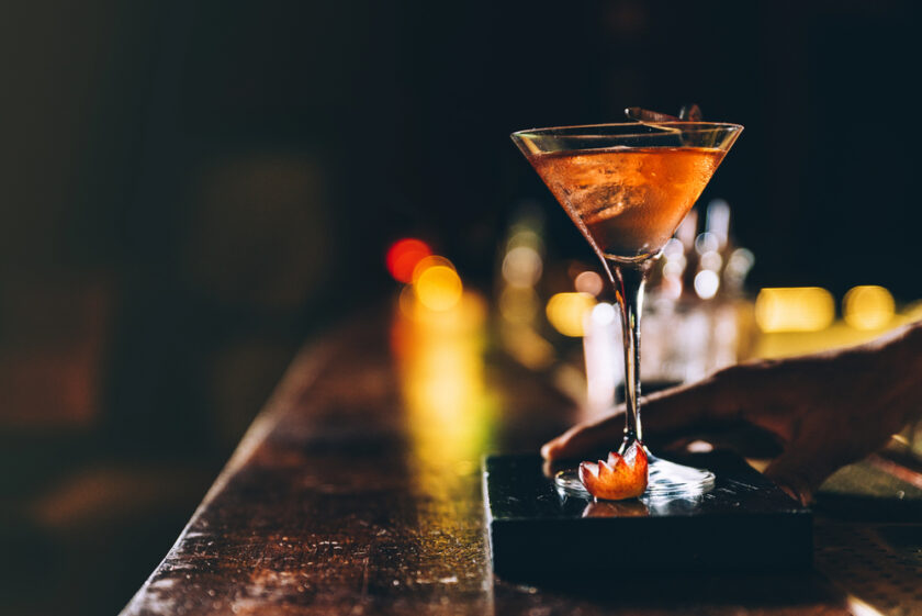 A hand reaches for a martini glass filled with an orange cocktail, garnished with a twist of citrus peel, sitting on a dark bar counter with a blurred background and warm, ambient lighting.