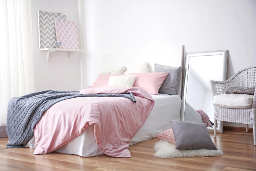 A cozy bedroom with a bed covered in pink and gray bedding, decorative pillows, a white chair, a mirror, and framed artwork on a shelf against a white wall, with wooden flooring.