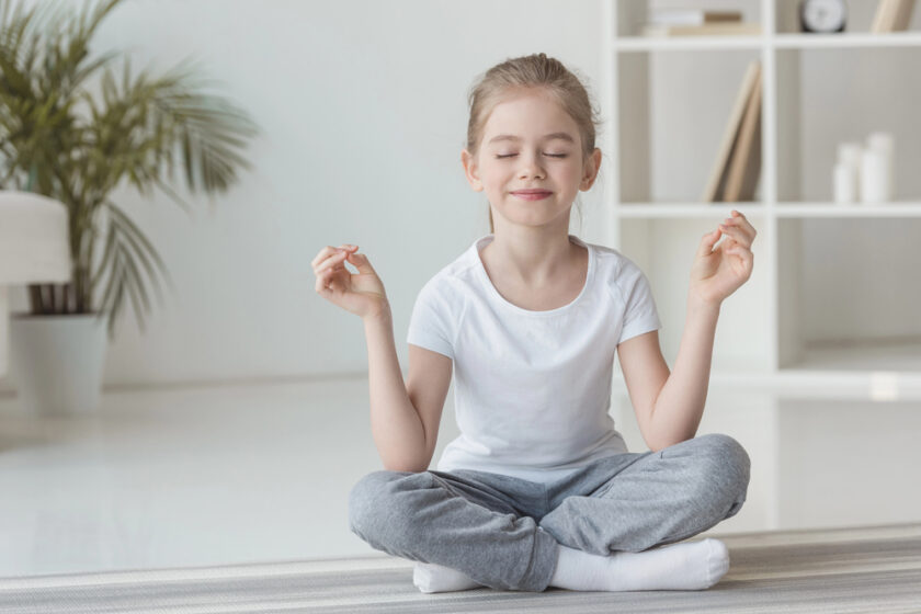A young girl sits cross-legged on a mat indoors, eyes closed and smiling, with her hands resting on her knees in a meditation pose. The background shows a plant and shelving unit.