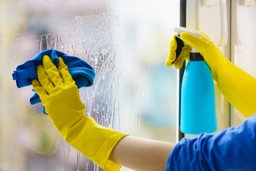 A person wearing yellow rubber gloves cleans a window with a blue cloth and sprays cleaning solution from a blue bottle.