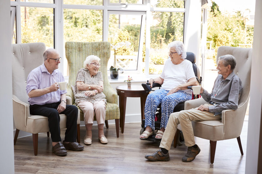 Four elderly people sit in armchairs by a window, smiling and chatting with each other. One woman uses a wheelchair. Two of them hold cups, and sunlight streams into the bright, cosy room.