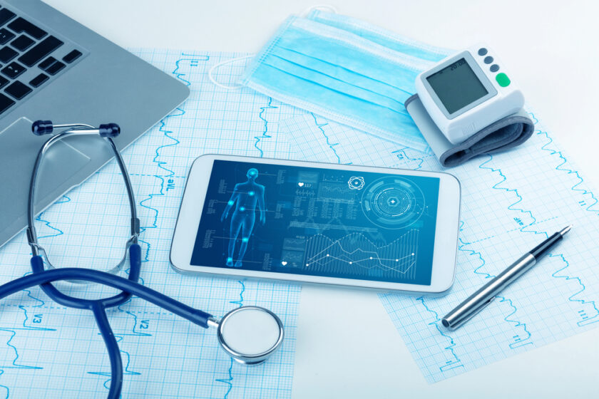A stethoscope, a tablet with medical data, a face mask, a digital blood pressure monitor, a pen, and printed charts are arranged on a desk next to a laptop.
