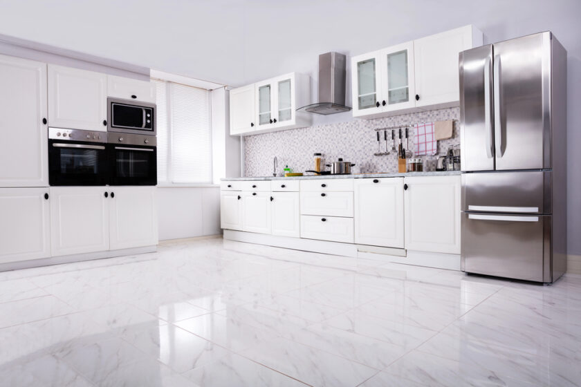 Modern kitchen with white cupboards, marble tiled floor, stainless steel fridge, built-in oven and microwave, and a splashback with small white tiles. Worktop holds various utensils and kitchen items.