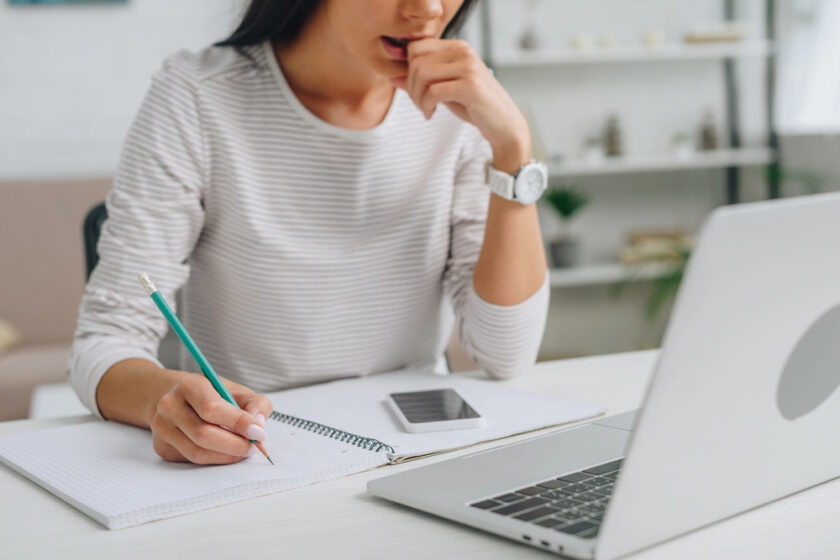 A woman in a striped shirt sits at a desk, writing in a notebook with a pencil whilst looking at a laptop. A mobile phone and more notebooks are on the desk. She appears focused and thoughtful.