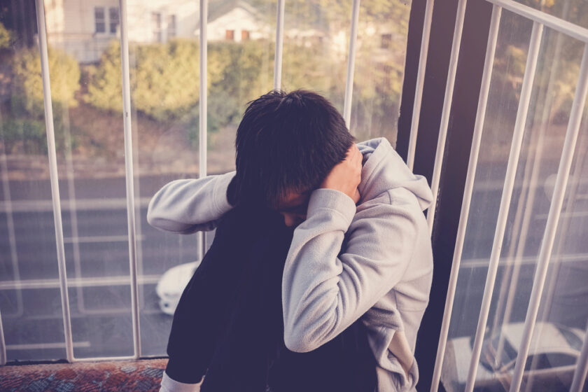 A person sits on the floor with their knees drawn up, head down, and hands covering their ears, appearing distressed. They are indoors near large windows overlooking a road and trees.