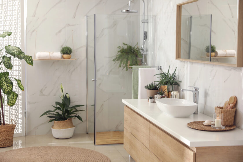 Modern bathroom with a glass shower, white marble walls, wooden vanity with basin, large mirror, potted plants, towels, candles, and natural light from a window.