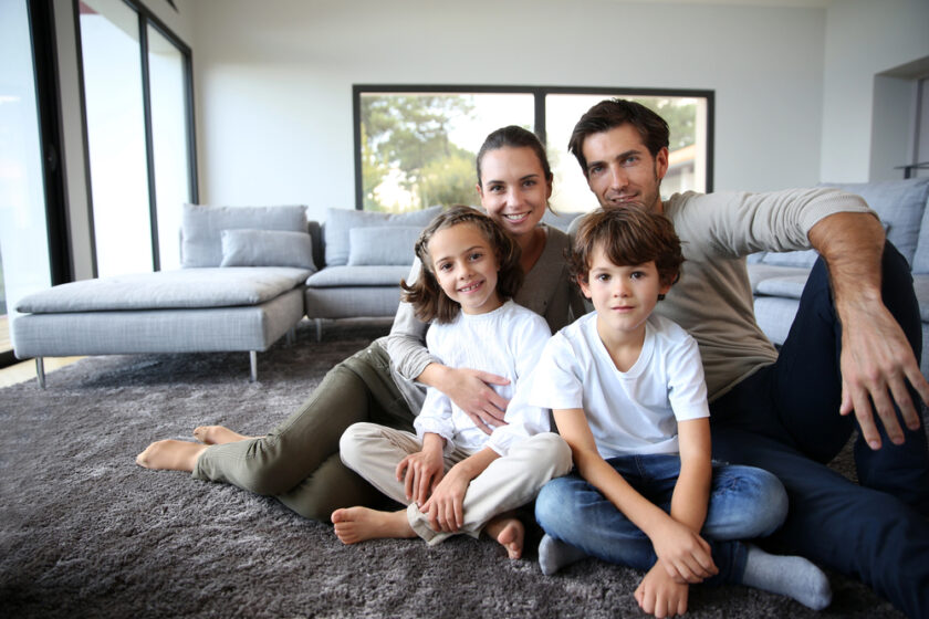 A smiling family of four sits together on a grey carpet in a bright, modern living room. Two children sit in front whilst their parents sit behind them, all looking at the camera.