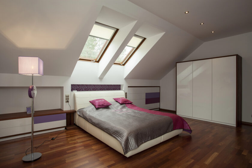 Modern attic bedroom with slanted ceiling, skylight windows, double bed with gray and purple bedding, nightstands, wardrobe, wooden floor, and a standing lamp. Natural light brightens the room.