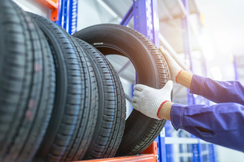 A person wearing white gloves places or removes a car tyre from a rack in a storage facility, with several other tyres lined up on the orange shelf.