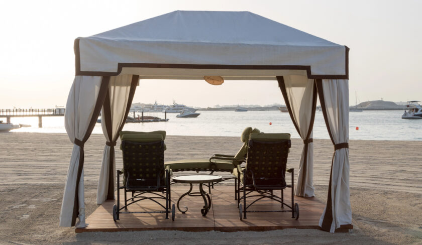 A white cabana with two loungers and a small table sits on a sandy beach, facing the water with boats in the distance. The scene is tranquil and sunlit.