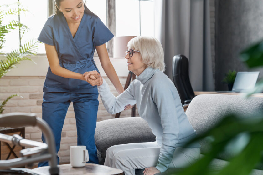 A nurse in blue scrubs helps an older woman with grey hair and glasses stand up from a sofa in a bright, cosy living room. A walking frame, coffee cup, and plants are visible in the background.